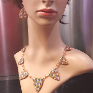 Può includere: Un set di collana e orecchini esposto su un manichino. I gioielli presentano ciondoli triangolari colorati con motivi a mosaico. La collana ha più ciondoli collegati da una catena d'argento e perline arancioni. Gli orecchini corrispondono al design della collana.