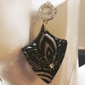 Può includere: Un singolo orecchino con un ciondolo nero a forma di diamante con un motivo floreale bianco. L'orecchino ha un perno color oro con un accento circolare tempestato di cristalli. Un piccolo cristallo trasparente è incastonato al centro del ciondolo.