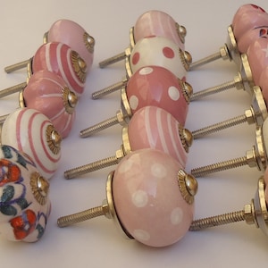 以下が含まれることがあります： ストライプ、水玉模様、花柄など、さまざまなパターンが施されたピンクのセラミック製キャビネットノブ12個。各ノブには銀色のネジが付いています。