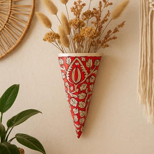 Op de afbeelding: Rode, kegelvormige wandvaas met een zilveren blad- en bloemmotief en een witte rand. De vaas, met gedroogde bloemen, is aan een neutrale muur bevestigd. Een wandkleed en een plant zijn ook zichtbaar.