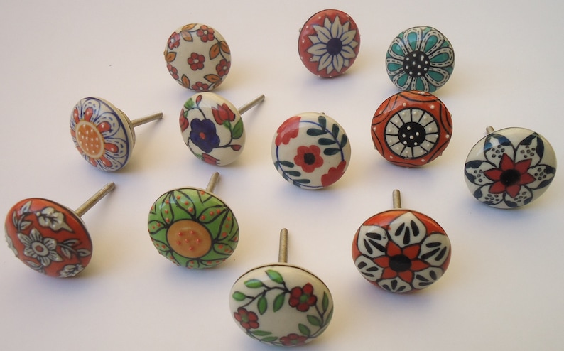 Pode incluir: Doze bot&otilde;es de cer&acirc;mica para gavetas com v&aacute;rios desenhos florais e geom&eacute;tricos. Os bot&otilde;es s&atilde;o pintados em tons de vermelho, laranja, verde, azul e branco.