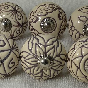 Peut inclure: Six boutons de meuble en c&eacute;ramique cr&egrave;me avec des motifs floraux et g&eacute;om&eacute;triques violets fonc&eacute;s. Chaque bouton a une base en m&eacute;tal argent&eacute;. Les boutons sont dispos&eacute;s en deux rang&eacute;es de trois.
