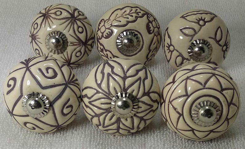 Peut inclure: Six boutons de meuble en c&eacute;ramique cr&egrave;me avec des motifs floraux et g&eacute;om&eacute;triques violets fonc&eacute;s. Chaque bouton a une base en m&eacute;tal argent&eacute;. Les boutons sont dispos&eacute;s en deux rang&eacute;es de trois.