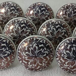 K&ouml;nnte beinhalten: Zehn wei&szlig;e Keramikkn&ouml;pfe mit einem schwarzen und kupferfarbenen Blumendesign. Die Kn&ouml;pfe sind rund und haben eine gl&auml;nzende Oberfl&auml;che.