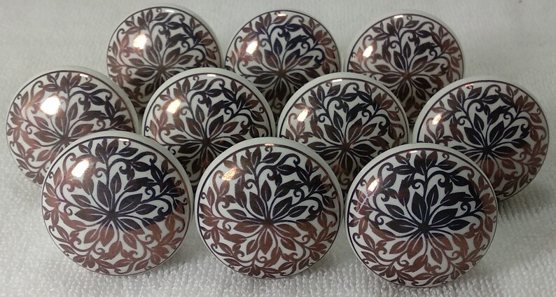 K&ouml;nnte beinhalten: Zehn wei&szlig;e Keramikkn&ouml;pfe mit einem schwarzen und kupferfarbenen Blumendesign. Die Kn&ouml;pfe sind rund und haben eine gl&auml;nzende Oberfl&auml;che.