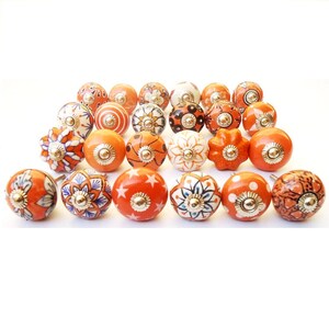 May include: A selection of orange and white ceramic cabinet knobs with diverse patterns. Designs include floral, star, and swirl motifs, each with a gold-toned metal base. The knobs are arranged on a white background.