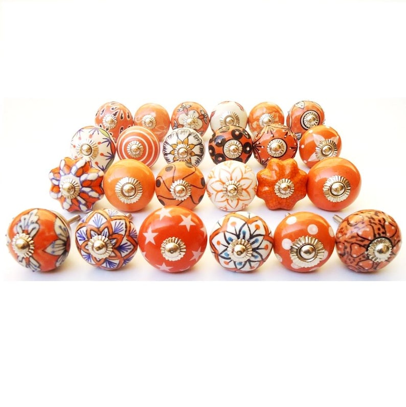 May include: A selection of orange and white ceramic cabinet knobs with diverse patterns. Designs include floral, star, and swirl motifs, each with a gold-toned metal base. The knobs are arranged on a white background.