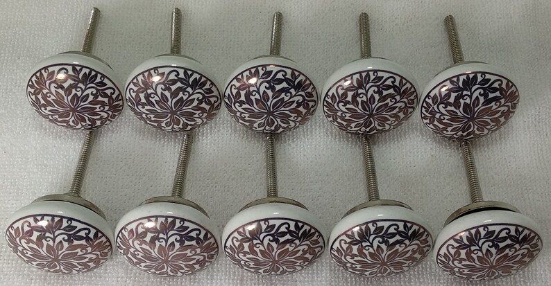 K&ouml;nnte beinhalten: Zehn wei&szlig;e Keramik-Schrankkn&ouml;pfe mit einer silbernen Schraube. Jeder Knopf hat ein braunes und wei&szlig;es Blumenmuster.