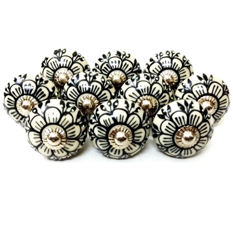 Peut inclure: Ensemble de boutons de placard en c&eacute;ramique cr&egrave;me avec un motif floral noir. Chaque bouton pr&eacute;sente un centre argent&eacute; et un motif floral d&eacute;coratif et stylis&eacute;. Les boutons sont dispos&eacute;s sur un fond blanc.