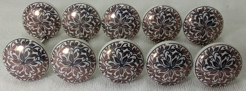 K&ouml;nnte beinhalten: Zehn wei&szlig;e Keramikkn&ouml;pfe mit einem schwarzen und kupferfarbenen Blumendesign. Die Kn&ouml;pfe haben ein erhabenes Design und eine glatte Oberfl&auml;che.