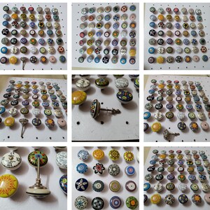 May include: A collection of colorful ceramic cabinet knobs with various floral and geometric designs. The knobs feature a range of colors, including blue, yellow, green, and red, and are mounted on a white surface. Some knobs have a silver metal base.