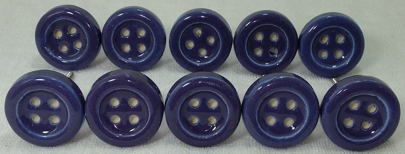 Op de afbeelding: Tien keramische knoppen in een diepe paarse kleur met een wit knoopontwerp. Elke knop heeft vier gaten in de knop.