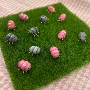 May include: A display of small, colourful woodlice figurines on a square of artificial green grass. The woodlice are painted in shades of pink, grey, and red with white legs. The background is a pink and white gingham pattern.