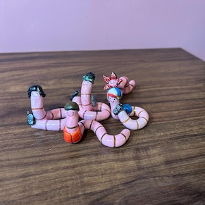 Op de afbeelding: Een verzameling grillige, roze wormfiguren met geschilderde details. Elke worm heeft een unieke uitdrukking en accessoires, waaronder hoeden en een kleine rugzak. De wormen zijn gerangschikt op een houten oppervlak.
