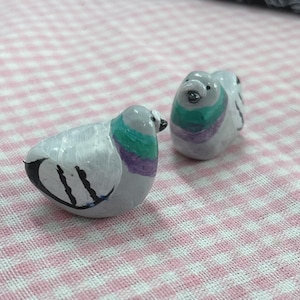 May include: Two small, hand-painted ceramic pigeon figurines. The birds are primarily white with grey heads, and feature teal, purple, and black accents. The figurines are on a pink and white checked surface.