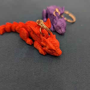 May include: Two small dragon keychains, one red and one purple, with gold rings. The red dragon is lying down with its head resting on the surface. The purple dragon is standing with its head tilted to the side.