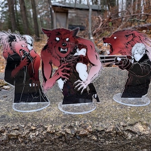 May include: Three acrylic standees featuring stylized illustrations of characters and a werewolf. The figures are in shades of red, black, and white, with clear bases. One figure is aiming a gun, while others are in a struggle.