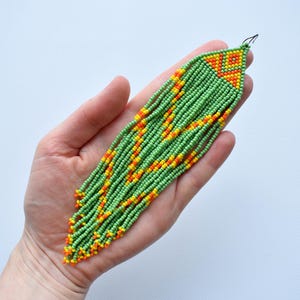 May include: A beaded earring with a fringe design. The earring is primarily green with orange and yellow accents. The earring is held in a hand against a white background. The earring is made of small beads.
