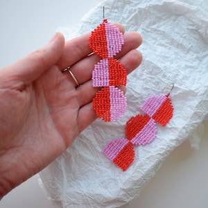 Può includere: Un paio di orecchini a forma di mezzaluna in perle rosa e rosse. Ogni orecchino è composto da tre forme a mezzaluna, con la metà superiore rosa e la metà inferiore rossa.