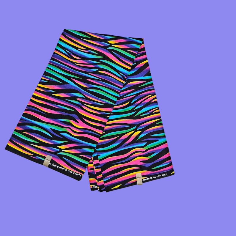 May include: A folded textile with a vibrant zebra stripe pattern in neon colors of pink, yellow, green, and blue on a black background. The fabric has the text "SUITABLE BLOCK WAX PRINTS" and "PRINCESS DUTCH WAX" on the edges.