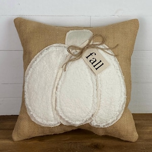 May include: A square throw pillow made of tan burlap fabric featuring a large, white, textured, sherpa-style pumpkin appliqué in the center. A small, rectangular, cream-colored tag tied with twine to the pumpkin stem displays the word fall.