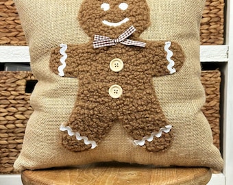 May include: A square throw pillow made of tan burlap fabric featuring a textured brown gingerbread man applique. The gingerbread man has white embroidered eyes and a smile, a brown and white checkered bow tie, two cream buttons, and white rickrack trim.