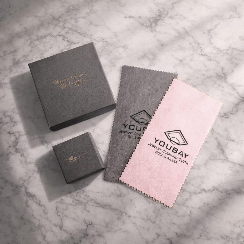 May include: Two dark gray square jewelry boxes with gold foil logos, one large and one small, accompanied by two rectangular cleaning cloths. One cloth is dark gray and the other is light pink, both featuring the black logo and text YOUBAY.