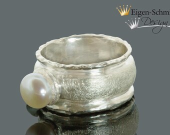 May include: A silver ring with a textured finish and a white pearl. The ring is wide and has a rounded shape.