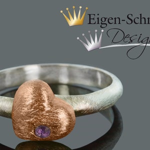 Puede incluir: Un anillo de plata con un engaste en forma de coraz&oacute;n de oro rosa con una peque&ntilde;a piedra preciosa p&uacute;rpura. El anillo est&aacute; sobre un fondo gris con el texto "Eigen-Schmied Design" encima.