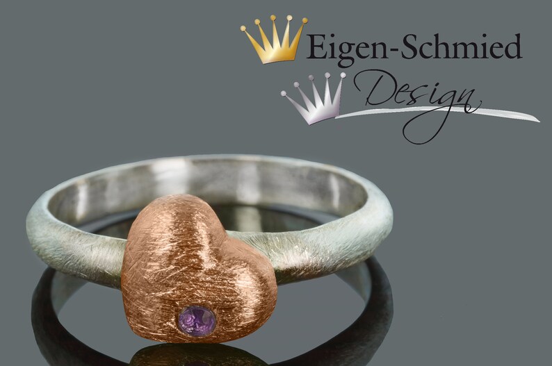 Puede incluir: Un anillo de plata con un engaste en forma de coraz&oacute;n de oro rosa con una peque&ntilde;a piedra preciosa p&uacute;rpura. El anillo est&aacute; sobre un fondo gris con el texto "Eigen-Schmied Design" encima.