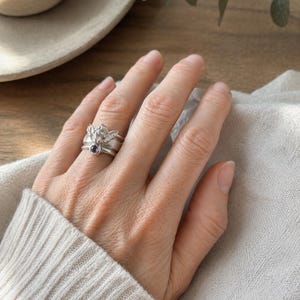 May include: A silver-toned ring featuring a detailed angel figure with wings spread wide, accented by a small, round, dark purple gemstone set at the base of the figure. The ring has a polished metallic finish and a smooth, rounded band design.