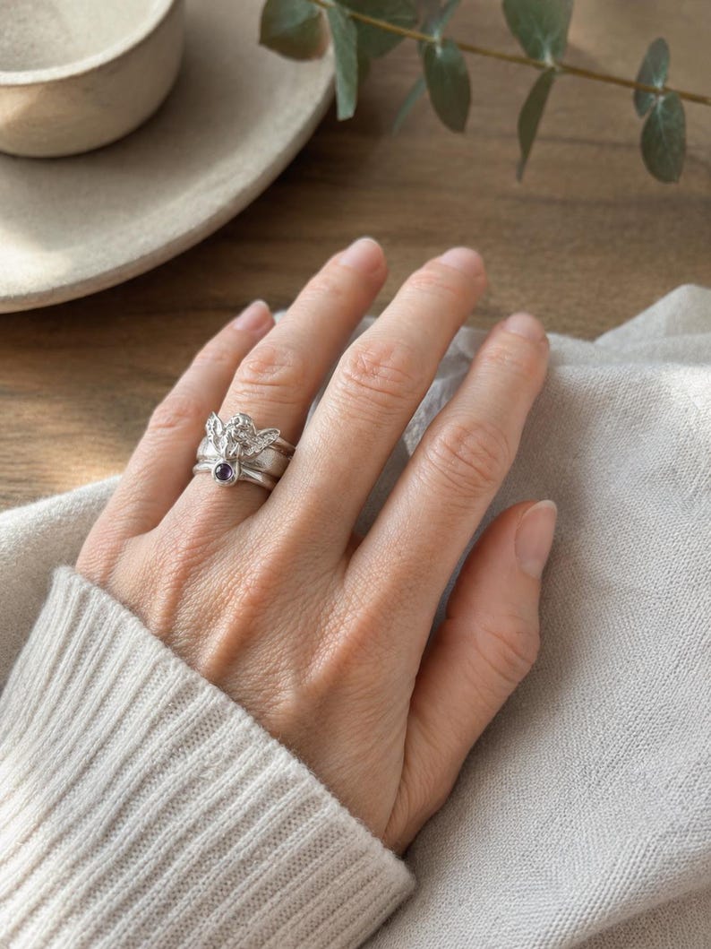 May include: A silver-toned ring featuring a detailed angel figure with wings spread wide, accented by a small, round, dark purple gemstone set at the base of the figure. The ring has a polished metallic finish and a smooth, rounded band design.