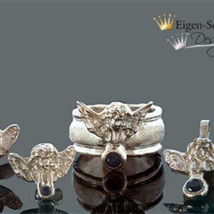 May include: A silver jewelry set featuring a ring and three matching angel-shaped charms, each adorned with a small dark blue gemstone. The ring displays a central angel figure with wings, and the text Eigen-Schmied Design appears in the corner.