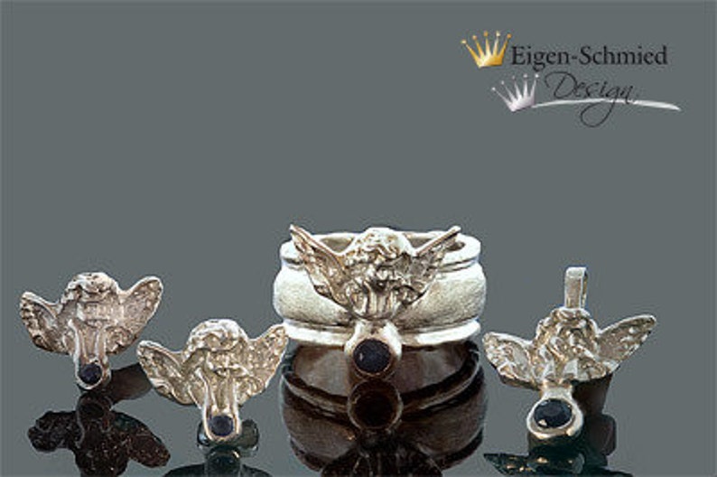 May include: A silver jewelry set featuring a ring and three matching angel-shaped charms, each adorned with a small dark blue gemstone. The ring displays a central angel figure with wings, and the text Eigen-Schmied Design appears in the corner.