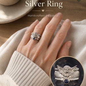 May include: A silver ring featuring a sculpted angel figure with wings spread wide and a small, round, dark blue gemstone set into the band. The text Guardian Angel Silver Ring, Handcrafted Elegance, and Symbol of Protection. Made to be with you. is visible.