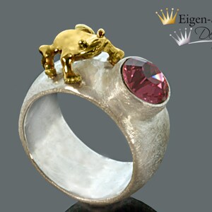 May include: A silver ring with a textured matte finish featuring a small gold-toned frog figure perched on top and a single round, faceted pink gemstone set into the band. The text Eigen-Schmied Design is visible in the upper right corner.