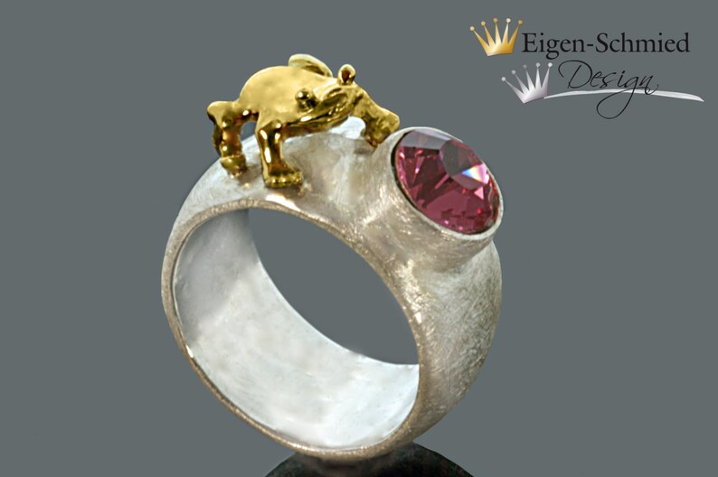 May include: A silver ring with a textured matte finish featuring a small gold-toned frog figure perched on top and a single round, faceted pink gemstone set into the band. The text Eigen-Schmied Design is visible in the upper right corner.