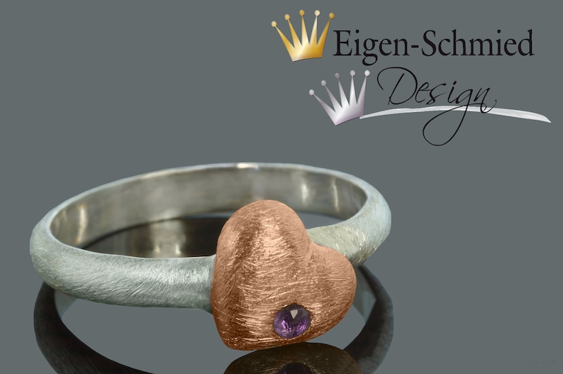 Puede incluir: Un anillo de plata con un engaste en forma de coraz&oacute;n de oro rosa y una peque&ntilde;a piedra preciosa p&uacute;rpura. El anillo est&aacute; sobre un fondo gris.