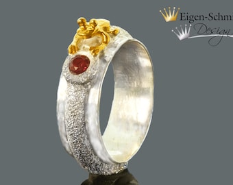 May include: A silver ring with a textured band and a gold frog figurine on top. The frog is holding a red gemstone.