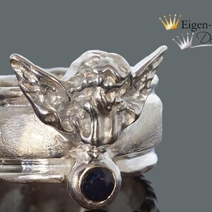May include: A silver ring featuring a sculpted angel figure with outstretched wings and a single round dark blue gemstone set at the base. The band has a textured finish and the text Eigen-Schmied Design is visible in the upper right corner.