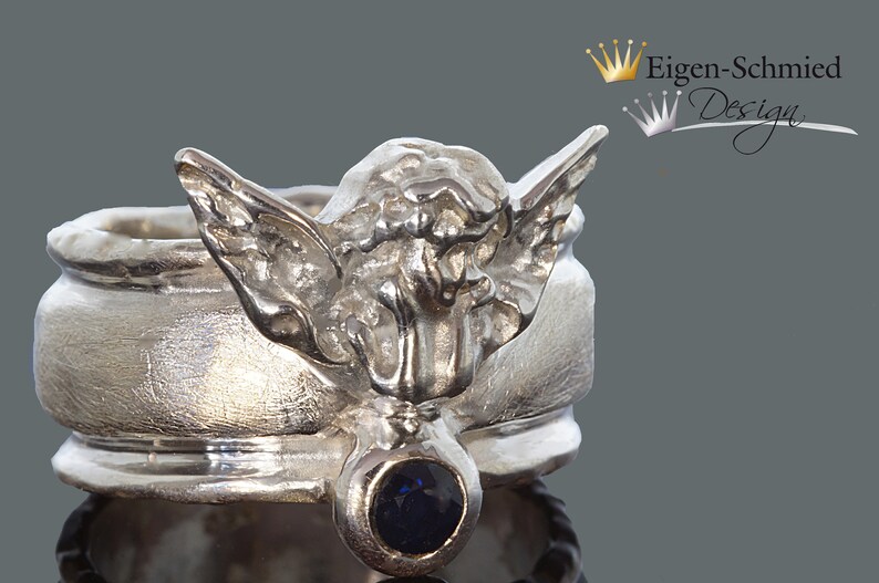 May include: A silver ring featuring a sculpted angel figure with outstretched wings and a single round dark blue gemstone set at the base. The band has a textured finish and the text Eigen-Schmied Design is visible in the upper right corner.