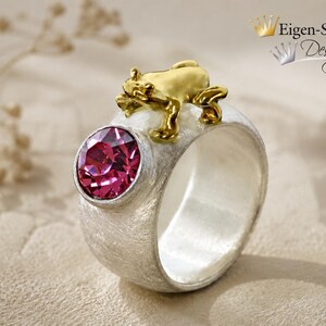 May include: A wide silver ring with a textured matte finish featuring a round, faceted fuchsia gemstone set in a bezel and a small, polished gold-toned frog figure perched on the top edge. The text Eigen-Schmied Design appears in the corner.