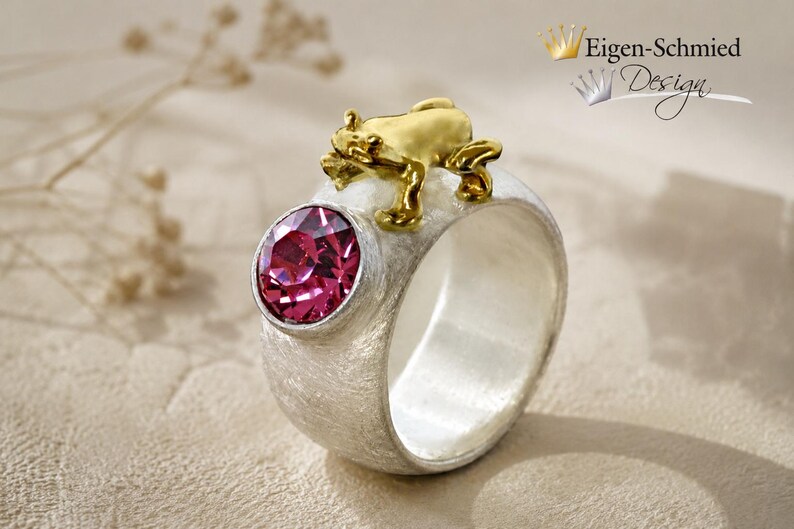 May include: A wide silver ring with a textured matte finish featuring a round, faceted fuchsia gemstone set in a bezel and a small, polished gold-toned frog figure perched on the top edge. The text Eigen-Schmied Design appears in the corner.