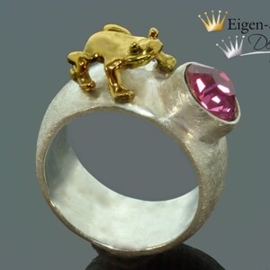 May include: A wide silver ring with a matte, textured finish featuring a small, polished gold-tone frog figure perched on top. A single, faceted, round pink gemstone is set into the band next to the frog. The text Eigen-Schmied Design is visible.