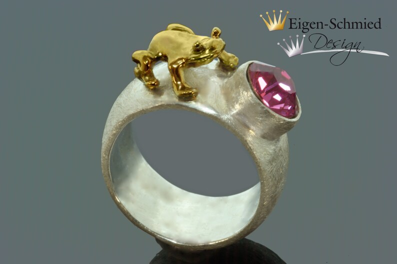 May include: A wide silver ring with a matte, textured finish featuring a small, polished gold-tone frog figure perched on top. A single, faceted, round pink gemstone is set into the band next to the frog. The text Eigen-Schmied Design is visible.