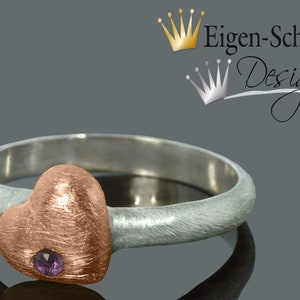 Puede incluir: Un anillo de plata con un engaste en forma de coraz&oacute;n de oro rosa con una peque&ntilde;a piedra preciosa p&uacute;rpura. El anillo est&aacute; hecho por Eigen-Schmied Design.