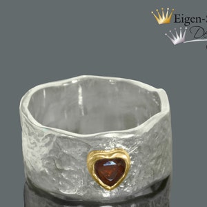 Könnte beinhalten: Ein silberner Ring mit strukturierter Oberfläche und einem goldfarbenen herzförmigen Akzent mit einem roten Edelstein. Der Ring hat ein breites Band und ein einzigartiges, handgefertigtes Aussehen. Es ist ein Schmuckstück.