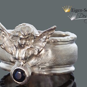 May include: A silver ring featuring a sculpted angel figure with outstretched wings and a single round dark blue gemstone set below the figure. The text Eigen-Schmied Design with a crown icon is visible in the upper right corner of the image.