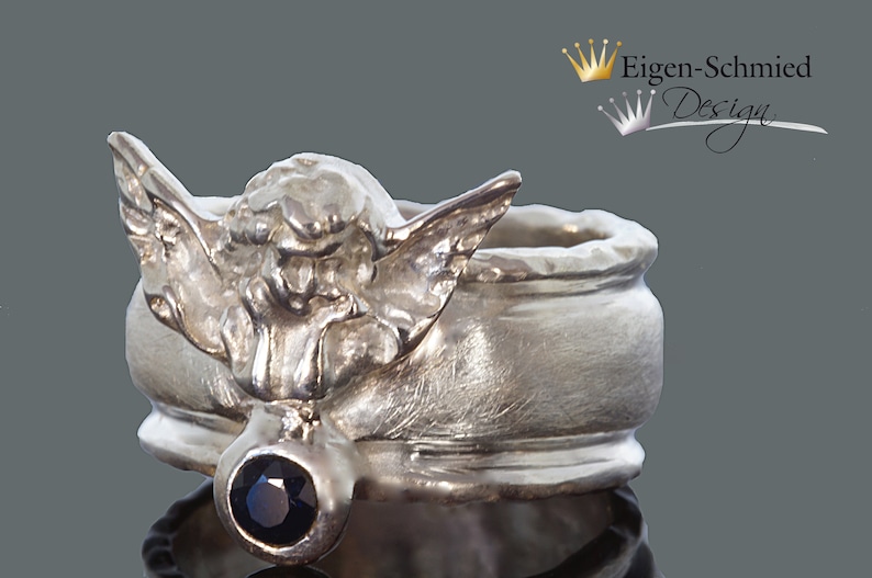 May include: A silver ring featuring a sculpted angel figure with outstretched wings and a single round dark blue gemstone set below the figure. The text Eigen-Schmied Design with a crown icon is visible in the upper right corner of the image.
