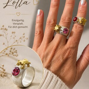 May include: Two silver rings with a textured finish, each featuring a gold-toned frog figure and a round faceted crystal. One ring has a pink crystal, and the other has a red crystal. The text Frosch Lotta, Einzigartig. Verspielt. F&uuml;r dich gemacht. 925 Silber & vergoldeter Frosch, Funkelnder Kristallstein, Mit Liebe handgefertigt, Eigen-Schmied Design is visible.